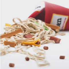 A red cone-shaped bag of dog treats tipped over on a white surface, with loose beige rope, brown chew pieces, and small yellow and green treats scattered in front.