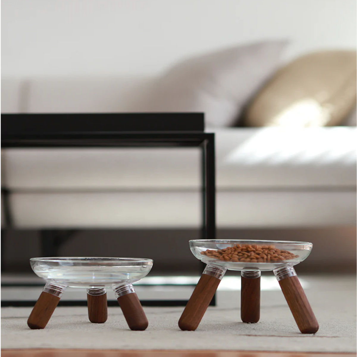 Two elevated pet bowls with clear glass tops and dark brown wooden legs, shown on a light rug in a softly blurred living room setting. One bowl is empty and the other contains kibble.