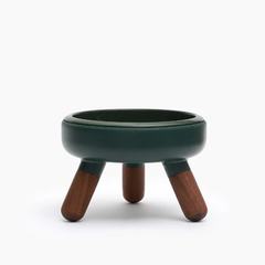A small round stool or planter with a dark green glazed ceramic bowl and three wooden legs, shown on a clean white background.