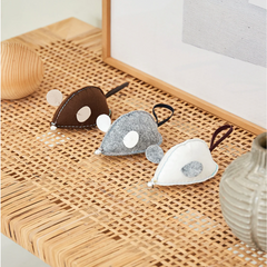 Three small felt mouse-shaped accessories in brown, gray, and white are arranged on a woven rattan surface with neutral decor in the background.